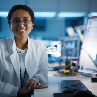 Female engineer wearing white lab coat sitting at desk with computers.