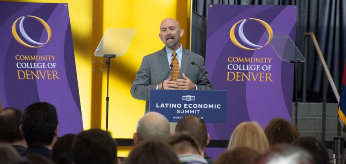 Photo of Joe Garcia speaking in front of a conference group with a microphone with signs behind him saying Community College of Denver.