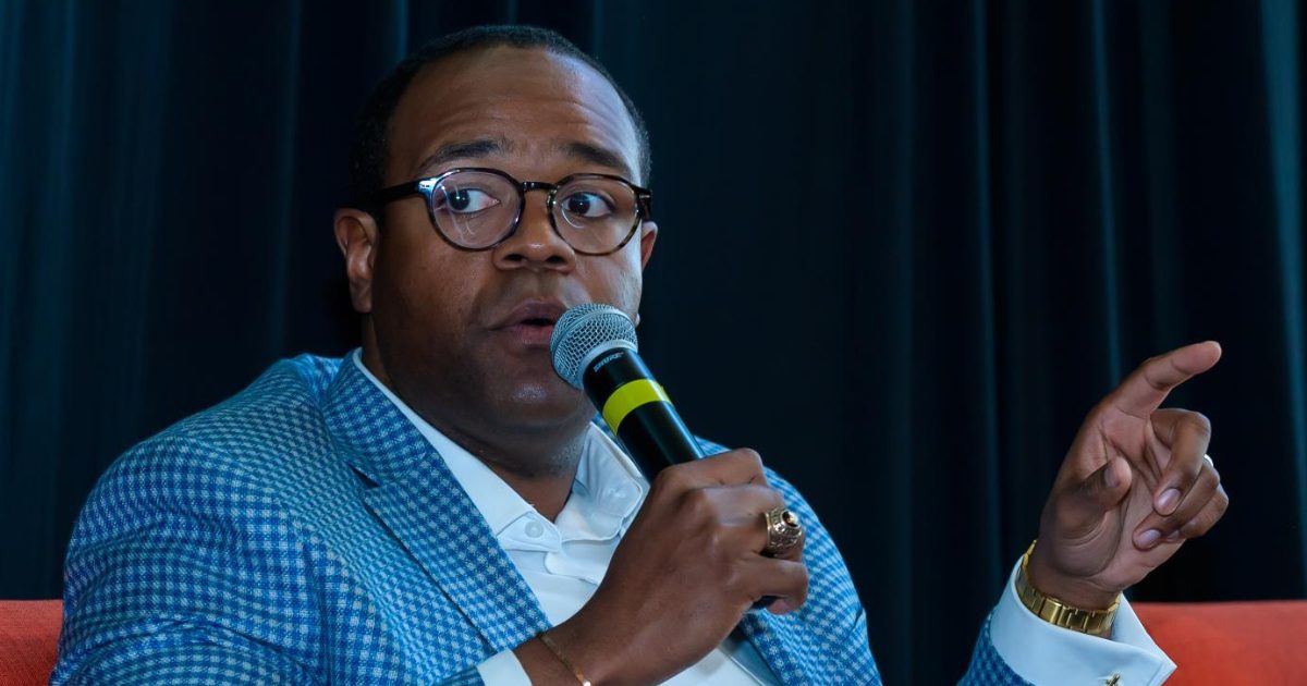 A man wearing glasses and a blue checkered blazer speaks into a microphone while gesturing with his hand.