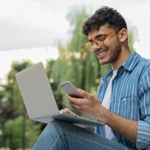 Young man with trendy glasses sitting outside with a laptop open smiling down at his cell phone.