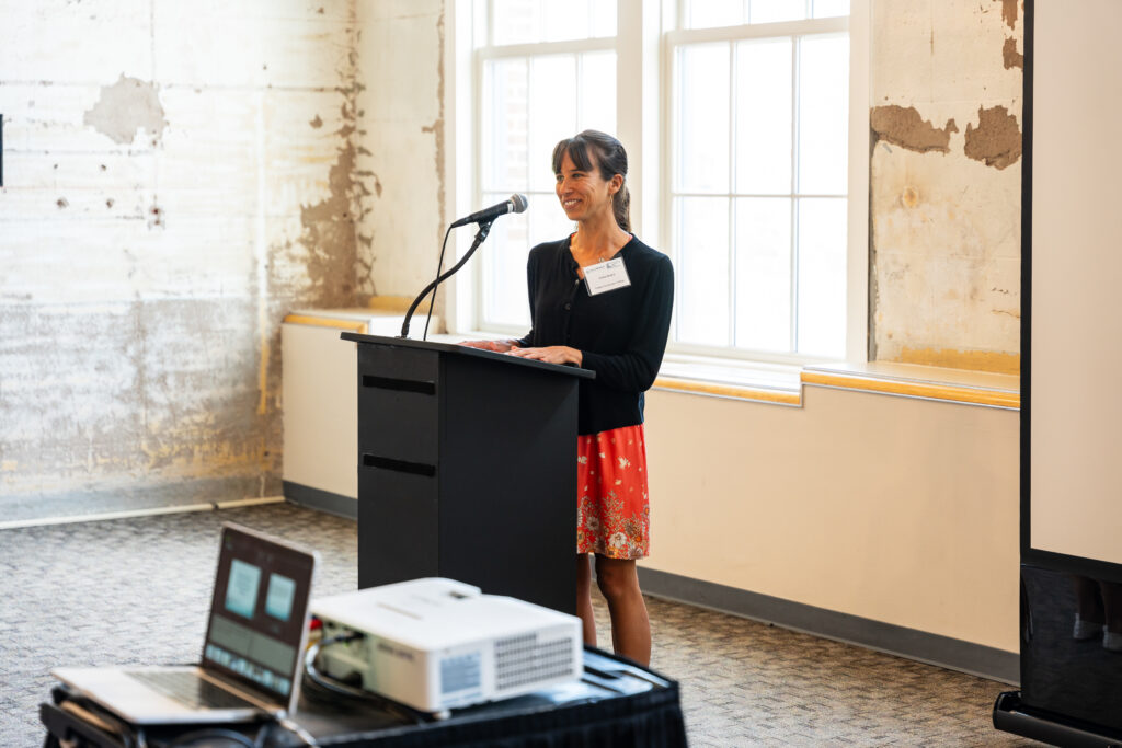 A woman stands at a podium with a microphone, smiling as she speaks. She is wearing a black cardigan and a red patterned skirt. Behind her is a wall with a weathered, textured appearance, and large windows letting in natural light. In the foreground, a laptop and projector are set up on a table.