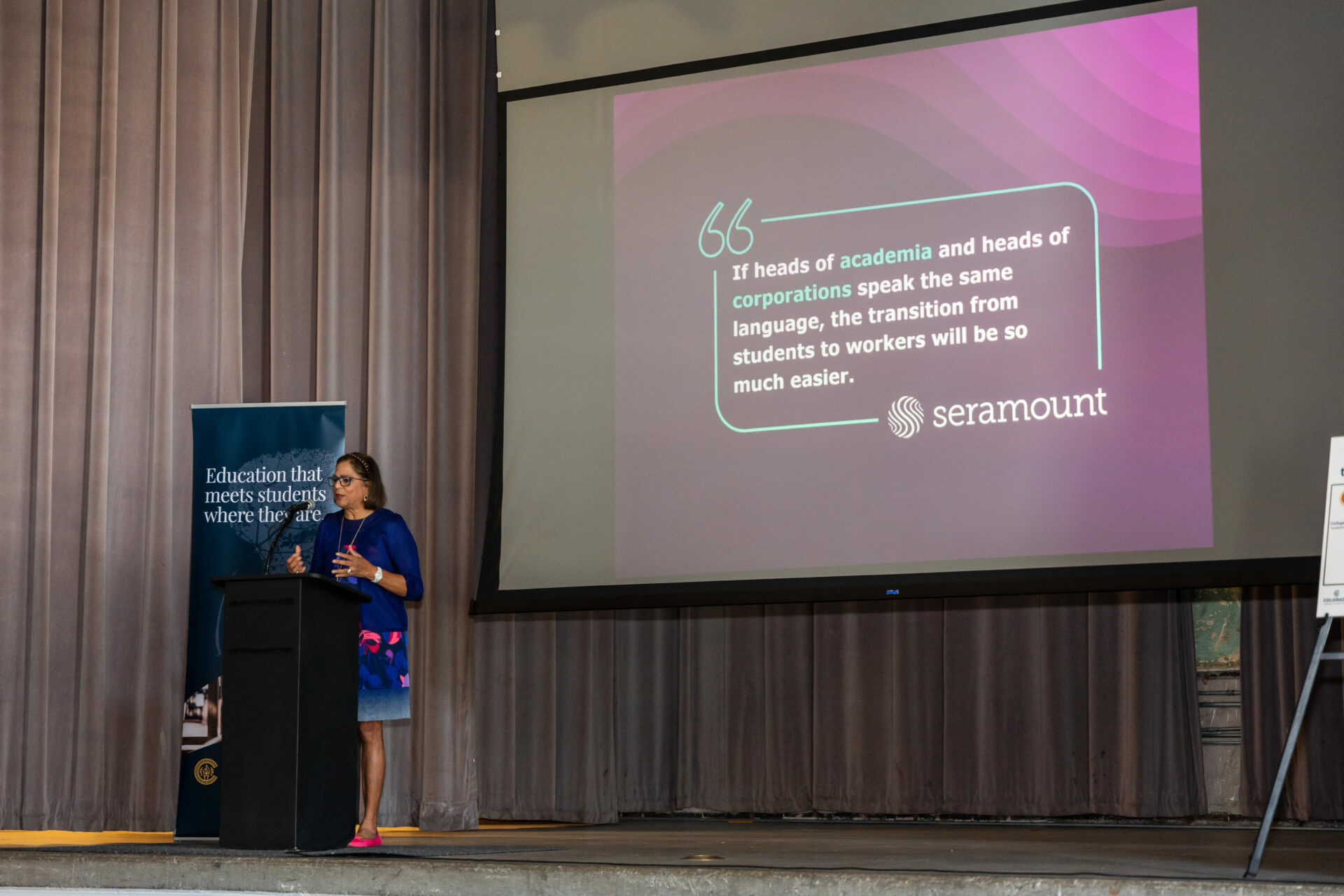 A speaker stands at a podium on a stage, delivering a presentation. Behind them, a large screen displays a quote about the importance of communication between academia and corporations for easing the transition from students to workers, attributed to 'seramount.' A banner to the left of the speaker reads, 'Education that meets students where they are.'