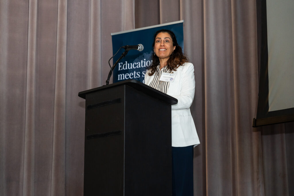 A woman stands at a podium with a microphone, smiling as she speaks. She is wearing a white blazer over a striped blouse. Behind her is a banner that reads, 'Education that meets students where they are.' The background consists of a curtain.