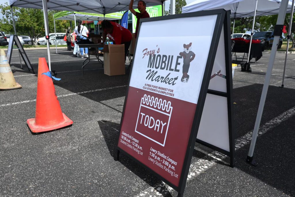 A sidewalk sign that reads "Foxy's Mobile Market," advertising a free food market for Community College of Aurora (CCA) students and employees. The market operates at two campus locations, listed on the sign, with a fox mascot illustration on the top right corner. Event tents and people setting up are visible in the background.