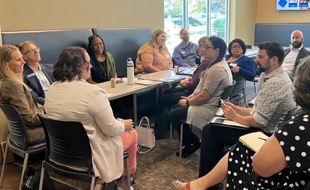 A diverse group of attendees sits in a casual breakout session setup, discussing topics in a relaxed environment. The group members face each other around a table, actively engaged in conversation.