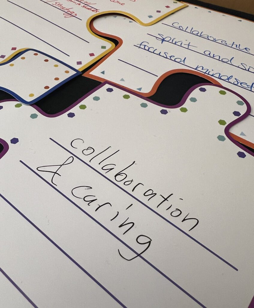 Close-up of a large, colorful puzzle piece with the handwritten words "Collaboration & Caring." The piece has multicolored dots around its edges and lines for additional text, symbolizing values or themes within the organization.